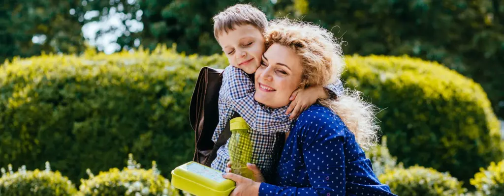 Eine Frau kniet und umarmt ihr Kind. Das Kind hat einen Schulrucksack auf dem Rücken. Die Frau hält eine Flasche und Brotdose fest.