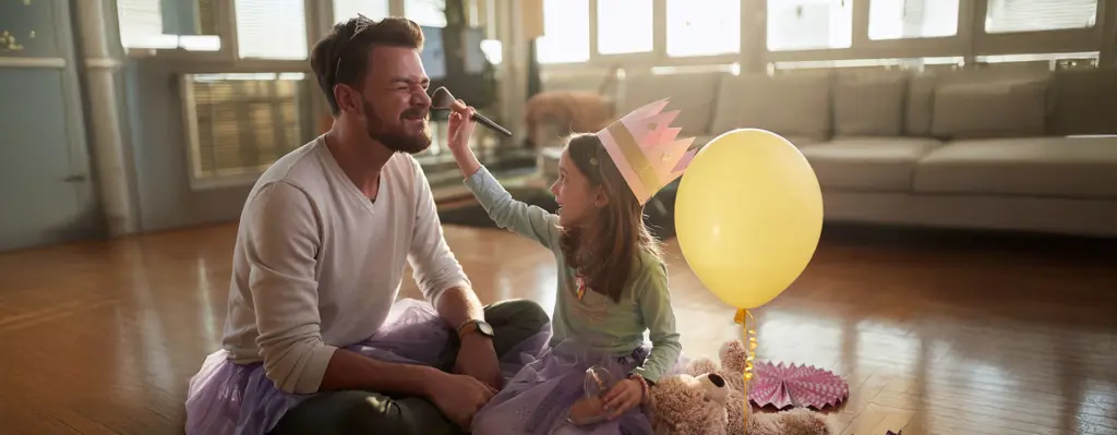 Vater sitzt im Tüllrock auf dem Boden, während seine Tochter ihm mit einem Pinsel Make-up aufträgt