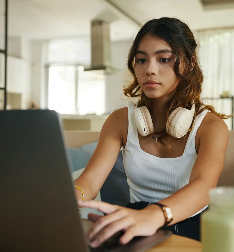 Junge Frau mit Kopfhörern um den Hals sitzt vor einem Laptop in einer Wohnung und tippt auf der Tastatur
