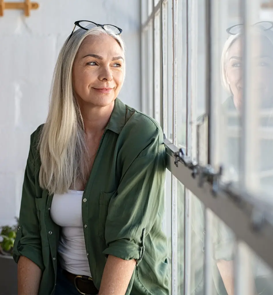 Frau mit langen grauen Haaren steht am Fenster und blickt nach draußen, ihr Spiegelbild ist im Glas zu sehen
