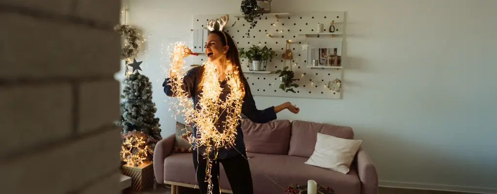 Frau steht im Wohnzimmer mit Lichterkette um den Hals und singt, im Hintergrund ist ein dekorierter Weihnachtsbaum
