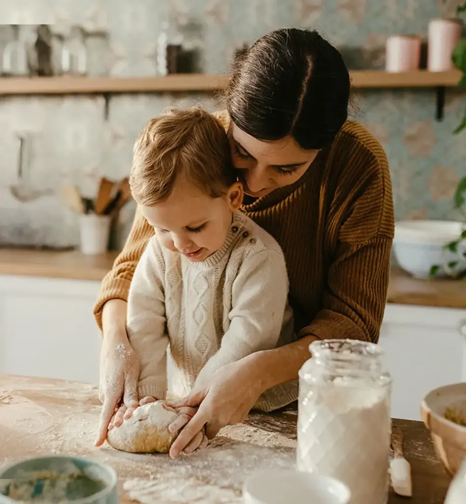 Mutter und Kleinkind kneten gemeinsam Teig auf einem bemehlten Holztisch in der Küche – innige, warme Stimmung beim Backen.