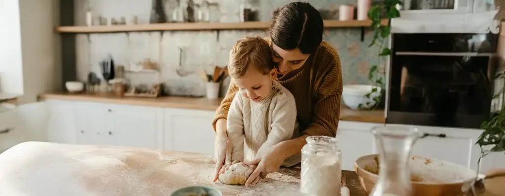 Mutter und Kleinkind kneten gemeinsam Teig auf einem bemehlten Holztisch in der Küche – innige, warme Stimmung beim Backen.