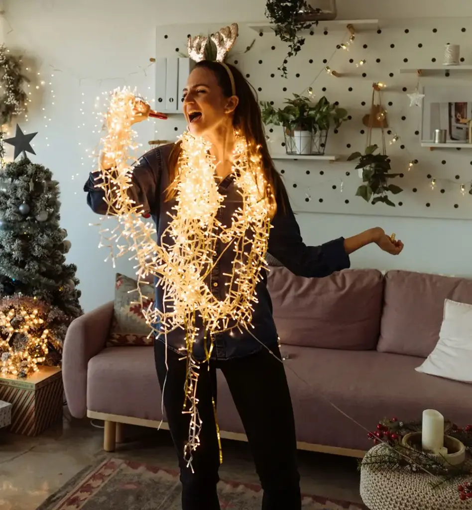 Frau mit Rentiergeweih-Haarreif tanzt ausgelassen im weihnachtlich dekorierten Wohnzimmer – in eine leuchtende Lichterkette gewickelt, singt sie fröhlich.