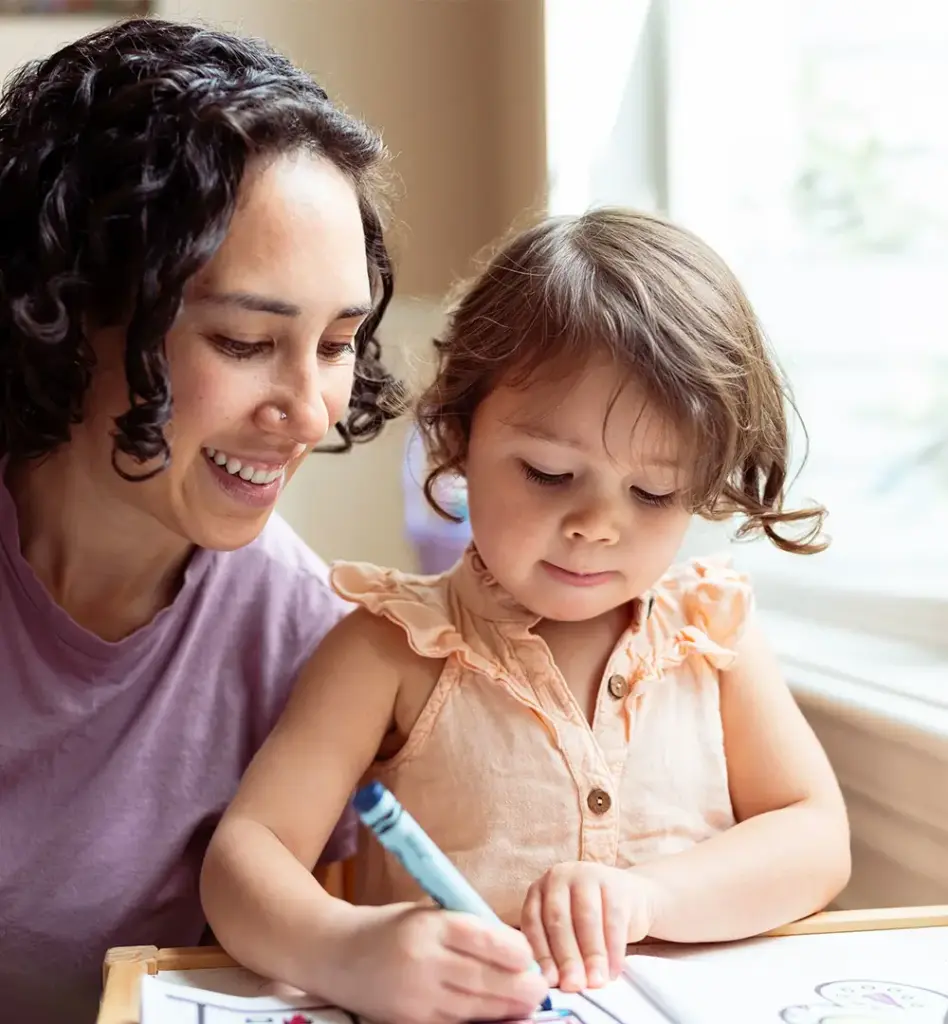Mutter schaut ihrer kleinen Tochter liebevoll beim Malen mit Buntstiften zu und lächelt dabei – helle, freundliche Atmosphäre.