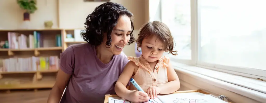 Mutter schaut ihrer kleinen Tochter liebevoll beim Malen mit Buntstiften zu und lächelt dabei – helle, freundliche Atmosphäre.