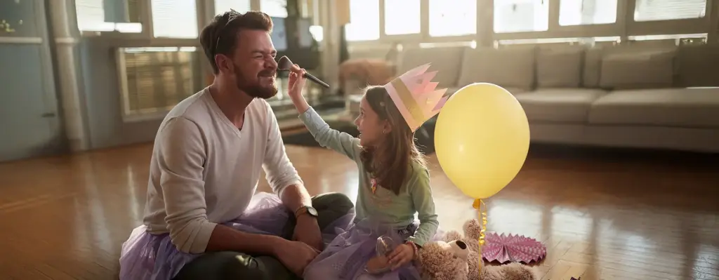 Vater und Tochter spielen verkleidet zuhause – Tochter mit Papierkrone schminkt den lachenden Vater mit einem Puderquast, beide tragen Tütüs.