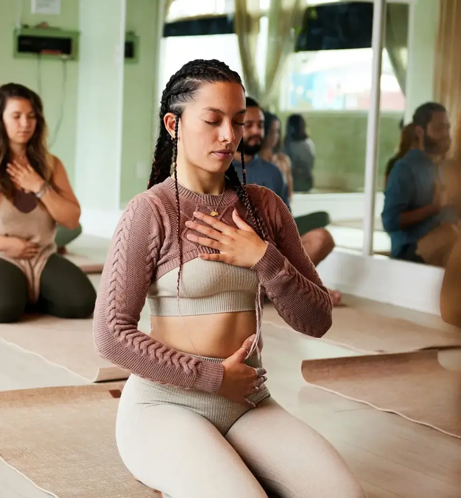 Frau mit Zöpfen sitzt im Schneidersitz auf einer Yogamatte und legt die Hände auf Herz und Bauch – konzentrierte Meditation in einer Yogagruppe.