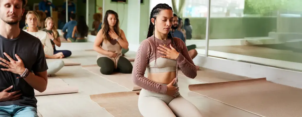 Frau mit Zöpfen sitzt im Schneidersitz auf einer Yogamatte und legt die Hände auf Herz und Bauch – konzentrierte Meditation in einer Yogagruppe.