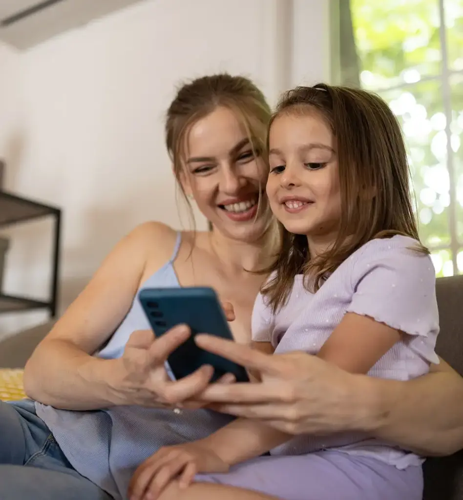 Mutter und Tochter schauen gemeinsam lächelnd auf ein Smartphone – sitzen entspannt auf einem Sofa, herzliche Stimmung.