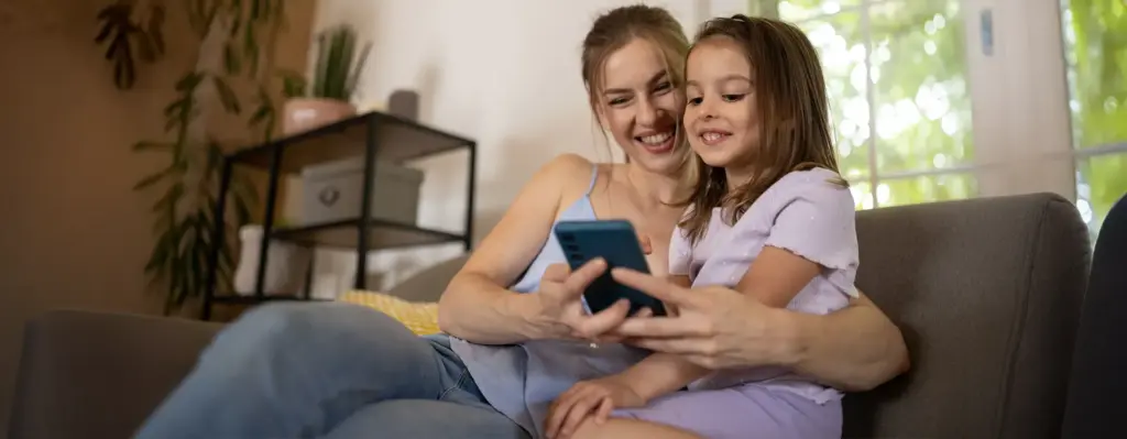 Mutter und Tochter schauen gemeinsam lächelnd auf ein Smartphone – sitzen entspannt auf einem Sofa, herzliche Stimmung.