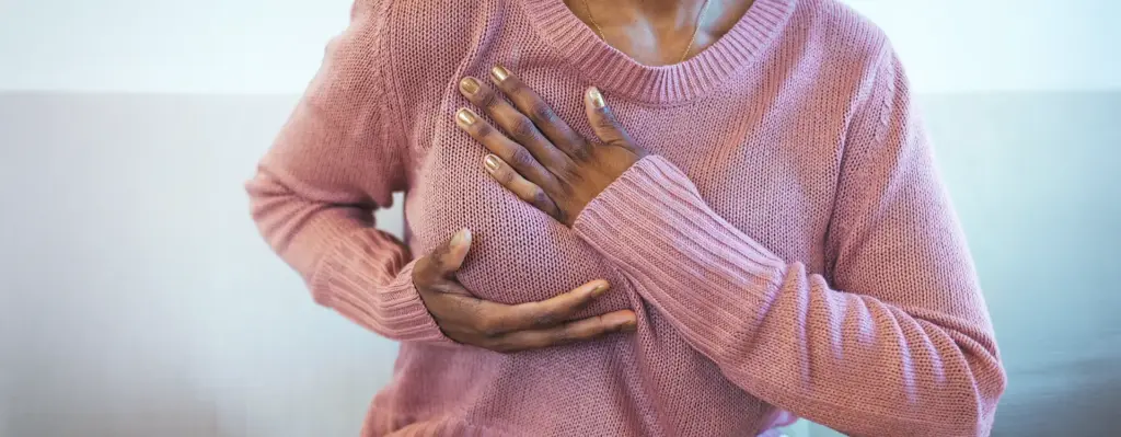 Nahaufnahme: Frau in rosa Pullover tastet ihre Brust mit beiden Händen ab – Eigenuntersuchung zur Brustkrebsvorsorge.
