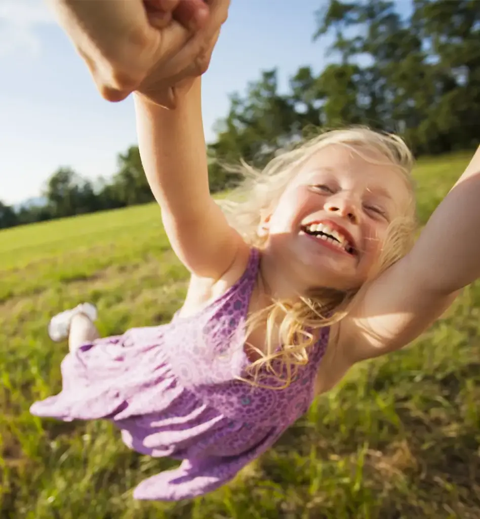 Lachendes blondes Mädchen im lila Kleid wird von einer erwachsenen Person an den Händen gehalten und ausgelassen im Kreis gedreht – sonniger Sommertag auf einer grünen Wiese.
