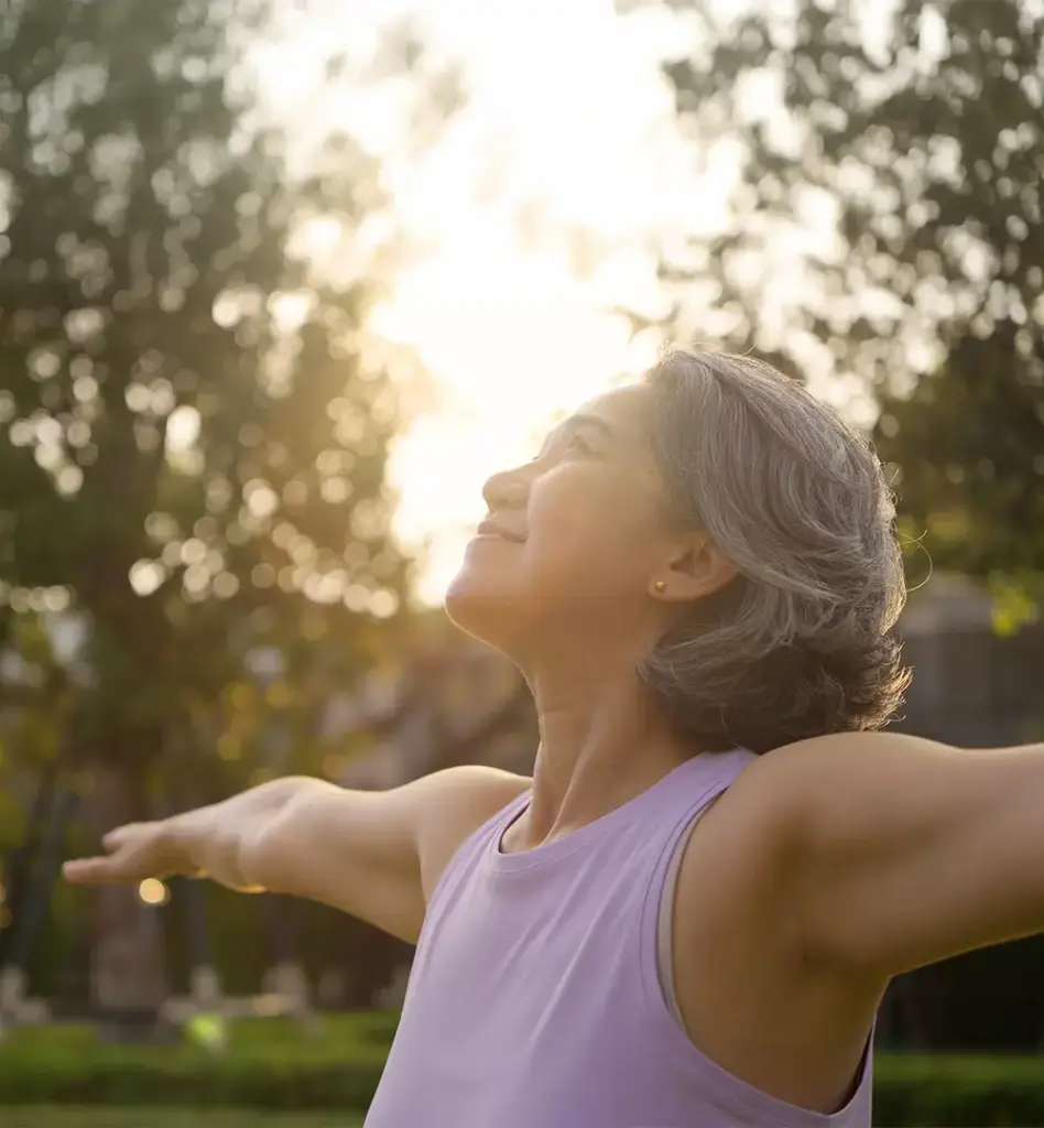 Ältere Frau mit grauem Haar und lila Sport-Top streckt die Arme aus und atmet tief durch – Outdoor-Workout im Park bei warmem Gegenlicht.