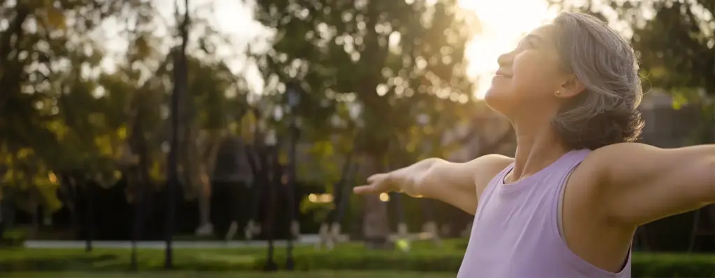 Ältere Frau mit grauem Haar und lila Sport-Top streckt die Arme aus und atmet tief durch – Outdoor-Workout im Park bei warmem Gegenlicht.