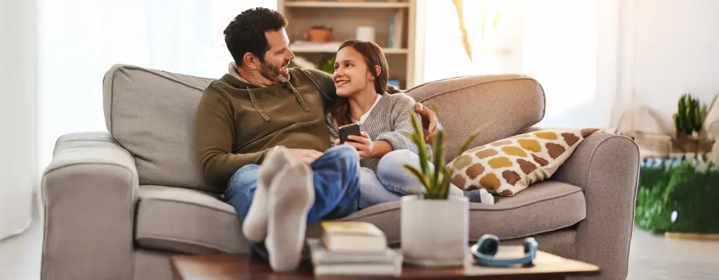 Vater und Teenager-Tochter sitzen entspannt auf dem Sofa und schauen sich lächelnd an – Tochter hält ein Smartphone in der Hand.