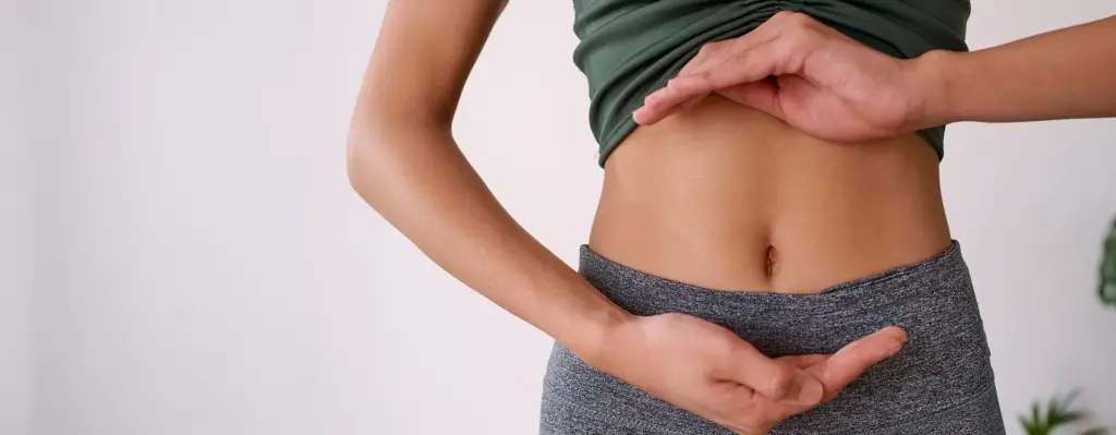 Nahaufnahme: Frau umrahmt mit beiden Händen ihren nackten Bauchbereich von oben und unten – seitliche Perspektive, neutraler Hintergrund.