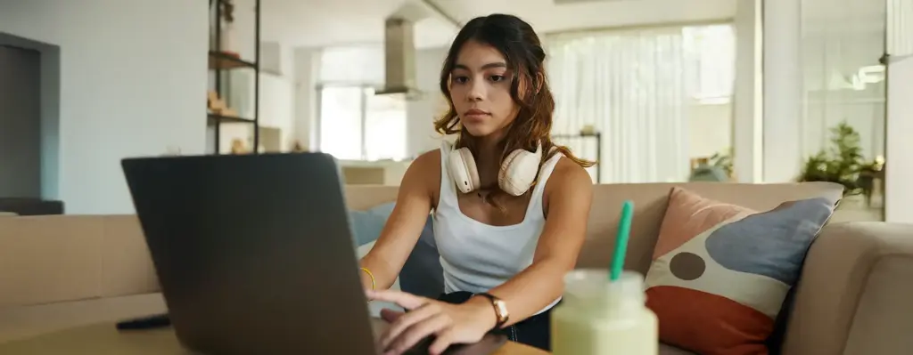 Junge Frau mit Kopfhörern um den Hals tippt konzentriert auf einem Laptop – helle, moderne Wohnatmosphäre im Hintergrund.