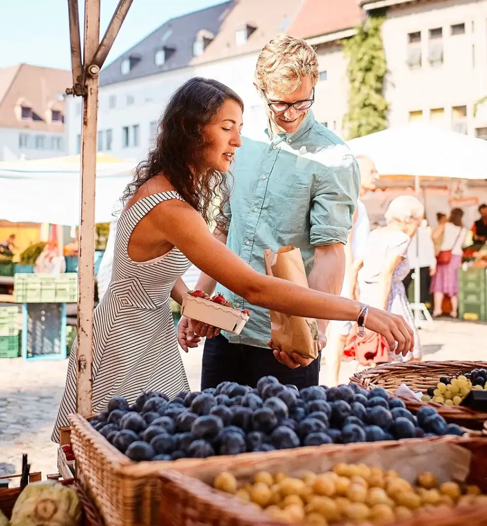 Paar kauft auf einem sommerlichen Wochenmarkt frisches Obst ein – Frau greift nach Pflaumen, Mann hält eine Papiertüte bereit.