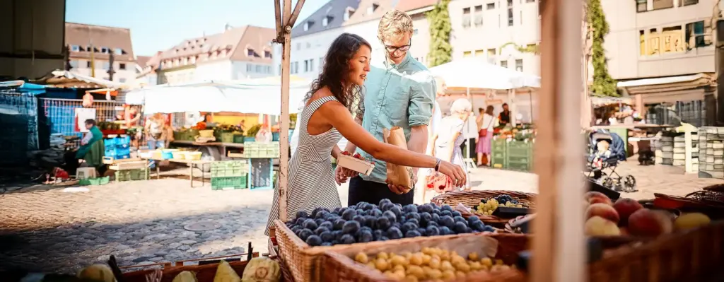 Paar kauft auf einem sommerlichen Wochenmarkt frisches Obst ein – Frau greift nach Pflaumen, Mann hält eine Papiertüte bereit.