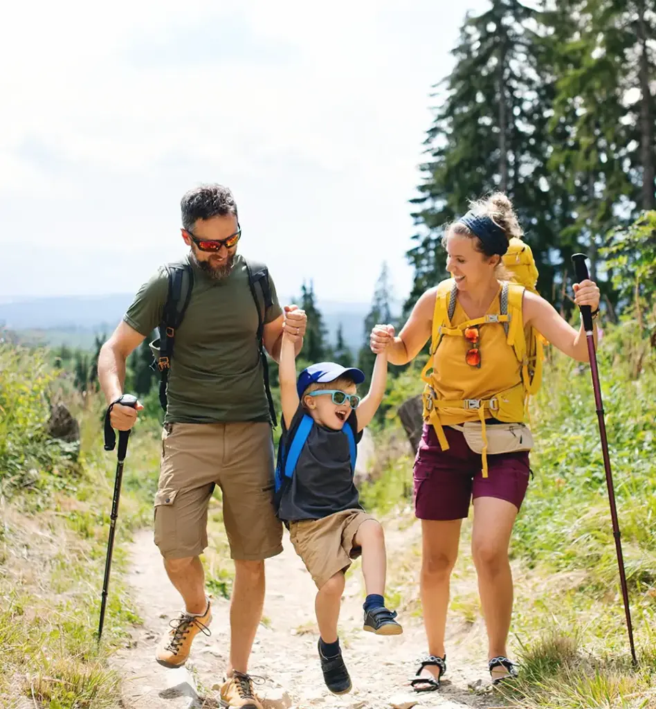 Familie wandert fröhlich auf einem Bergpfad – Eltern mit Rucksäcken und Wanderstöcken halten ihr lachendes Kind an den Händen und heben es hoch.