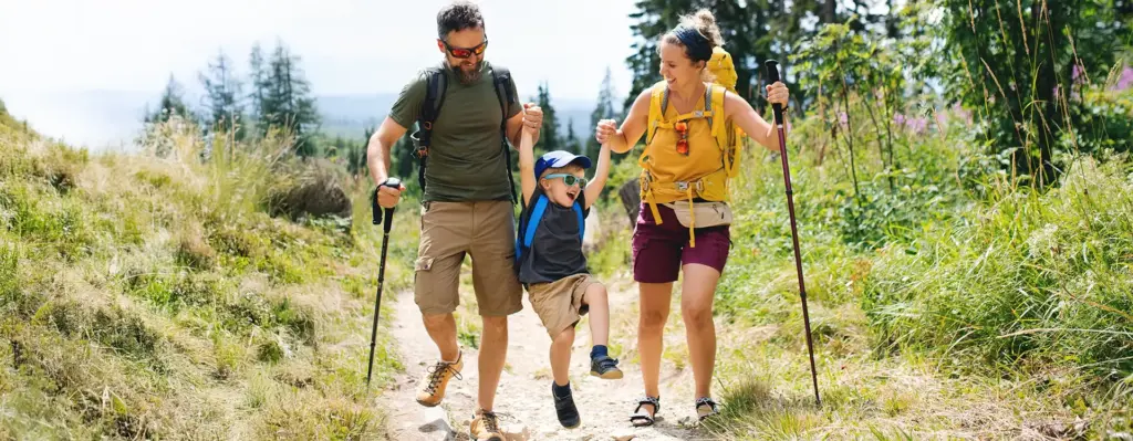 Familie wandert fröhlich auf einem Bergpfad – Eltern mit Rucksäcken und Wanderstöcken halten ihr lachendes Kind an den Händen und heben es hoch.
