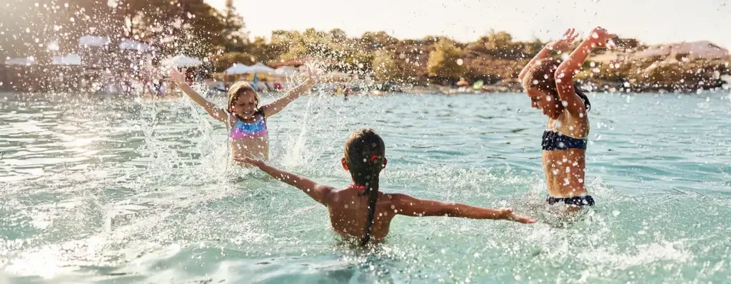 Drei Kinder spielen ausgelassen im türkisblauen Wasser – spritzen sich gegenseitig nass und jubeln.