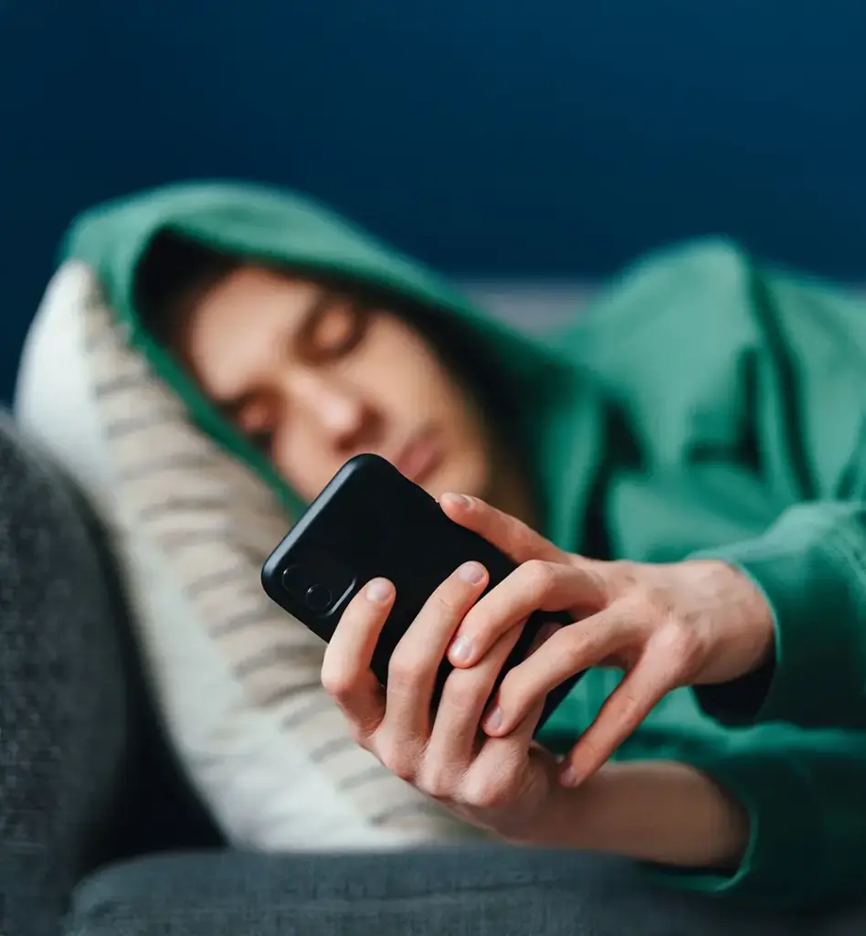 Teenager liegt erschöpft auf einem Sofa und scrollt mit halbgeschlossenen Augen auf seinem Smartphone – grüner Hoodie, dunkler Hintergrund.