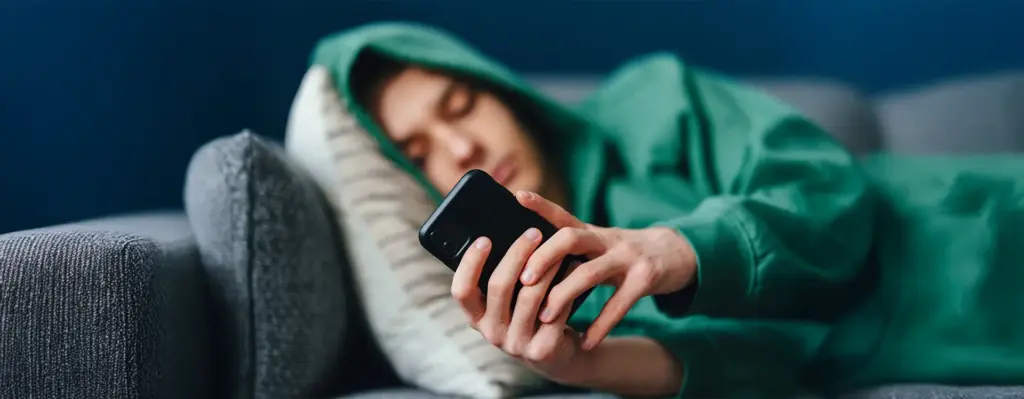 Teenager liegt erschöpft auf einem Sofa und scrollt mit halbgeschlossenen Augen auf seinem Smartphone – grüner Hoodie, dunkler Hintergrund.
