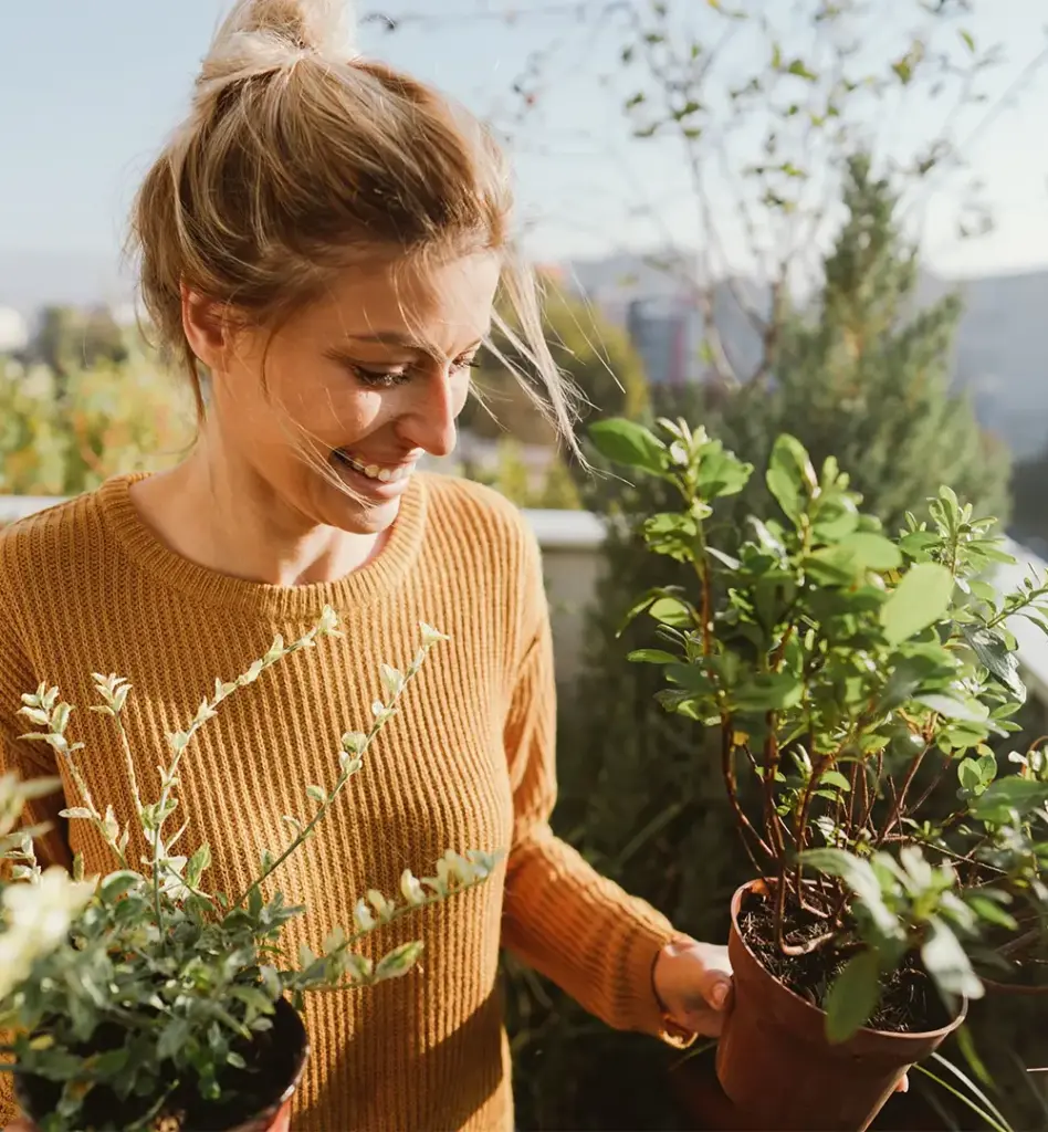 Junge Frau hält lächelnd zwei Topfpflanzen auf einem sonnigen Balkon – Stadtpanorama im Hintergrund, herbstliche Stimmung.