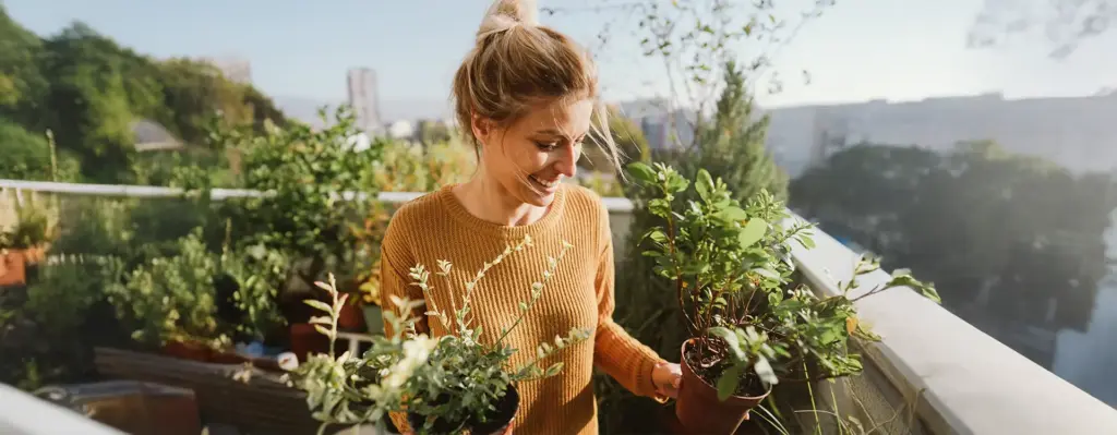 Junge Frau hält lächelnd zwei Topfpflanzen auf einem sonnigen Balkon – Stadtpanorama im Hintergrund, herbstliche Stimmung.