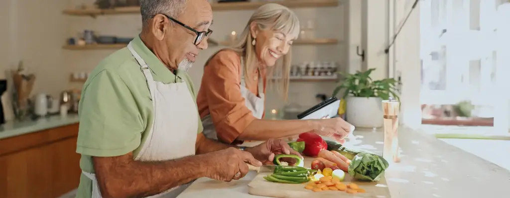 Älteres Paar kocht gemeinsam in der Küche – Mann schneidet Paprika, Frau lächelt und schaut auf ein Tablet mit Rezept.