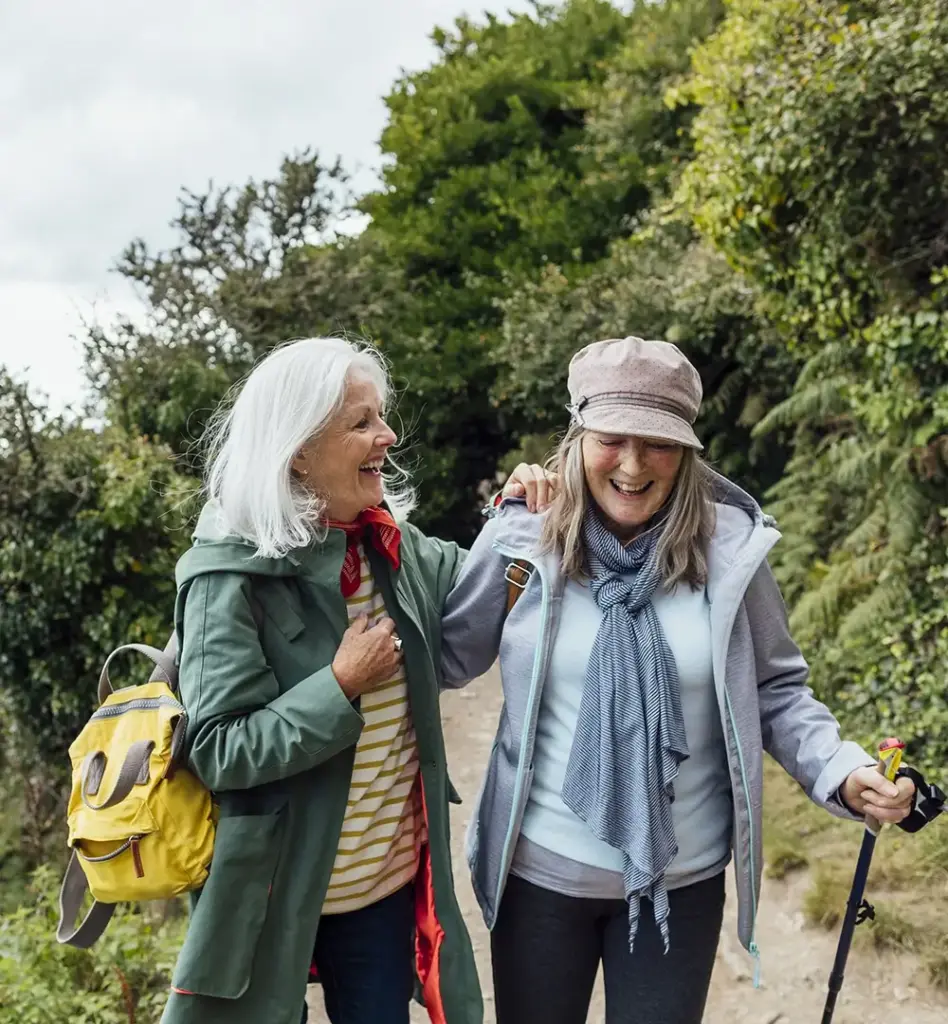 Zwei ältere Frauen wandern lachend Arm in Arm auf einem Naturpfad – fröhliche Stimmung, grüne Umgebung.