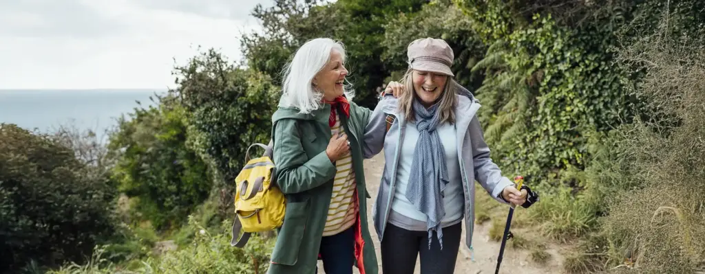 Zwei ältere Frauen wandern lachend Arm in Arm auf einem Naturpfad – fröhliche Stimmung, grüne Umgebung.