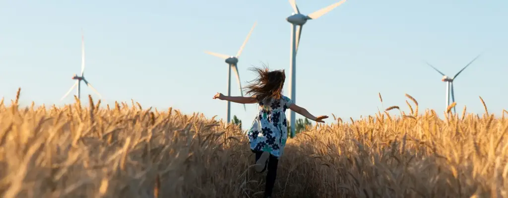 Mädchen rennt mit ausgebreiteten Armen durch ein goldenes Kornfeld – im Hintergrund zwei große Windkrafträder vor blauem Himmel.