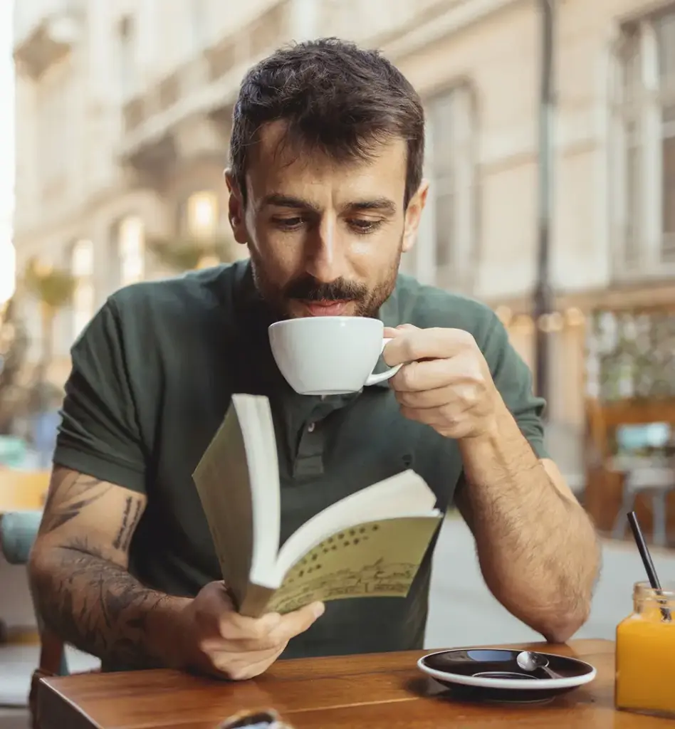 Mann trinkt Kaffee und liest ein Buch im Straßencafé – tätowierter Arm, entspannte Stimmung in urbaner Umgebung.