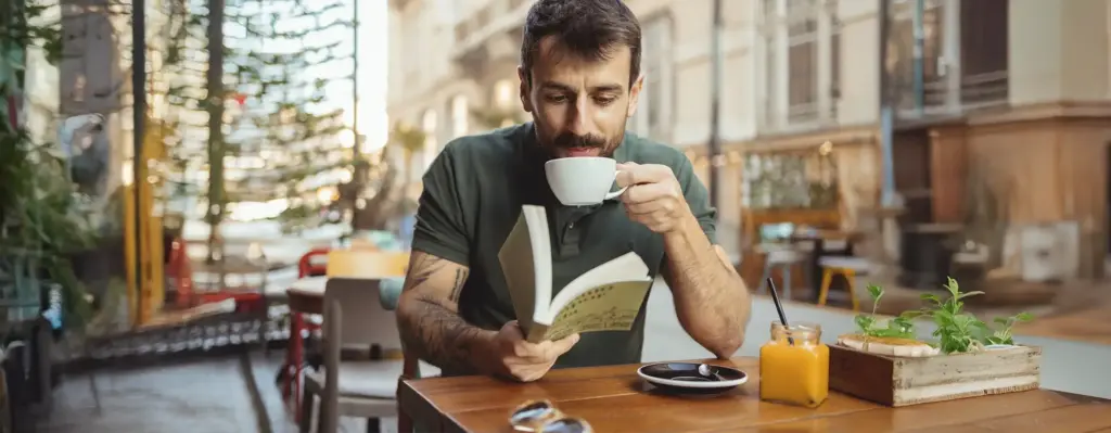 Mann trinkt Kaffee und liest ein Buch im Straßencafé – tätowierter Arm, entspannte Stimmung in urbaner Umgebung.