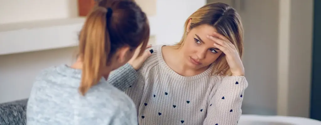 Zwei junge Frauen sitzen zusammen – eine legt der anderen fürsorglich die Hand auf die Schulter, während die andere besorgt wirkt und sich die Hand vor die Stirn hält.