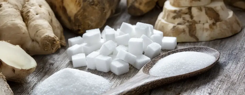 Zuckerwürfel und ein Holzlöffel mit Kristallzucker auf einem rustikalen Holztisch – aufgeschnittene Zuckerrüben im Hintergrund.