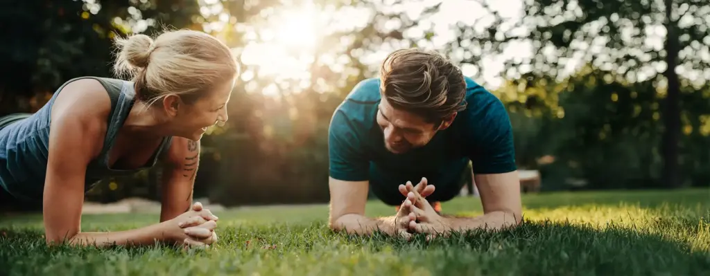 Paar macht gemeinsam Plank-Übungen auf einer Wiese im Park – schauen sich lächelnd an, warmes Gegenlicht der Abendsonne.