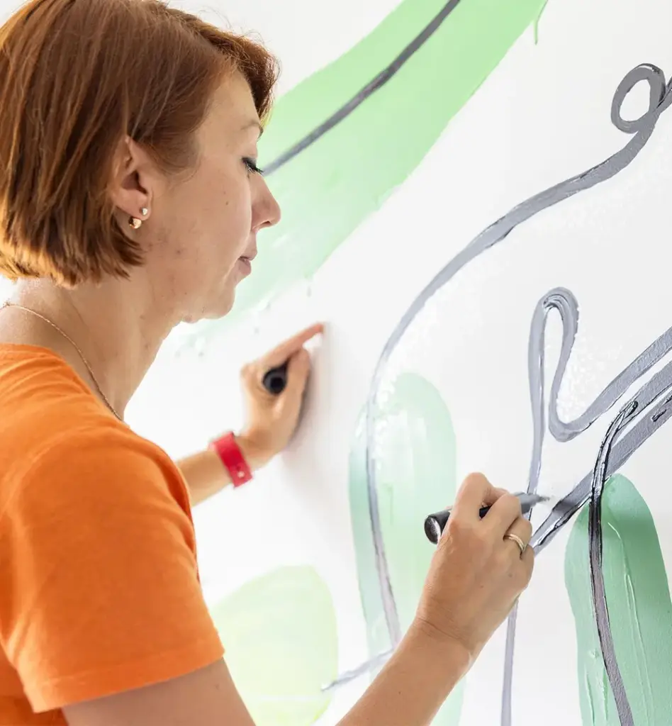 Frau in orangefarbenem T-Shirt malt konzentriert mit zwei Pinseln abstrakte Motive in Grün und Grau auf eine weiße Wand.