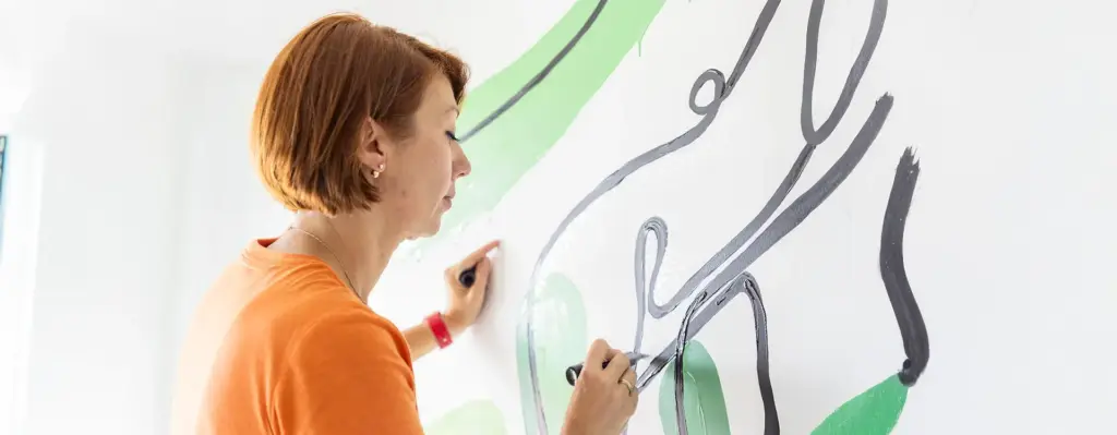 Frau in orangefarbenem T-Shirt malt konzentriert mit zwei Pinseln abstrakte Motive in Grün und Grau auf eine weiße Wand.