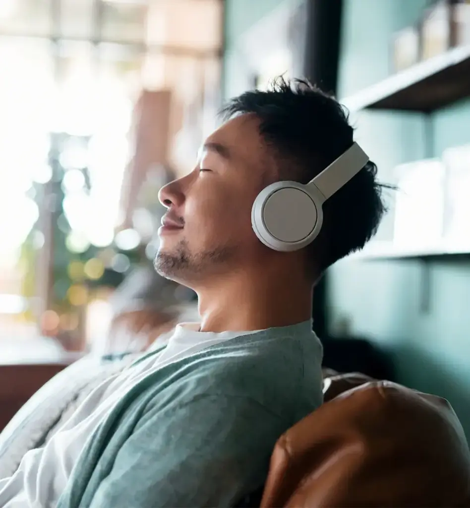 Junger Mann mit Kopfhörern lehnt entspannt auf einem Sofa und genießt Musik mit geschlossenen Augen – helle, ruhige Wohnatmosphäre.
