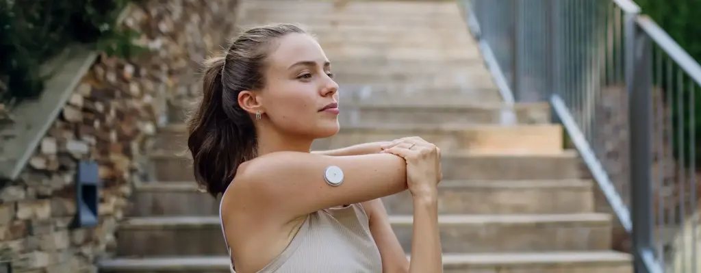 Junge Frau streckt sich vor dem Laufen – trägt am Oberarm einen CGM-Glukosesensor und an der Hüfte eine Insulinpumpe, aktives Leben mit Diabetes.