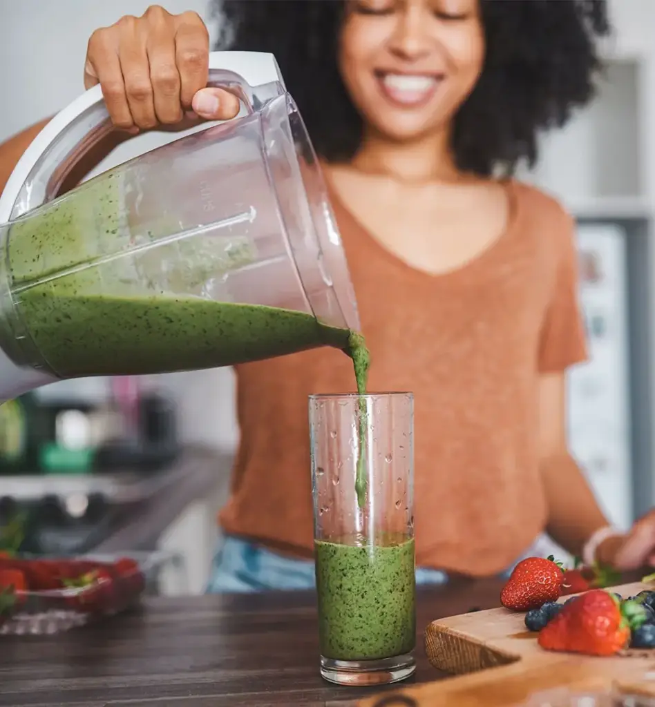 Lächelnde Frau mit Afro-Haar gießt einen grünen Smoothie aus dem Standmixer in ein hohes Glas – frische Erdbeeren und Beeren auf der Küchentheke.