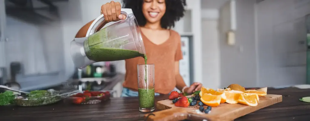 Lächelnde Frau mit Afro-Haar gießt einen grünen Smoothie aus dem Standmixer in ein hohes Glas – frische Erdbeeren und Beeren auf der Küchentheke.