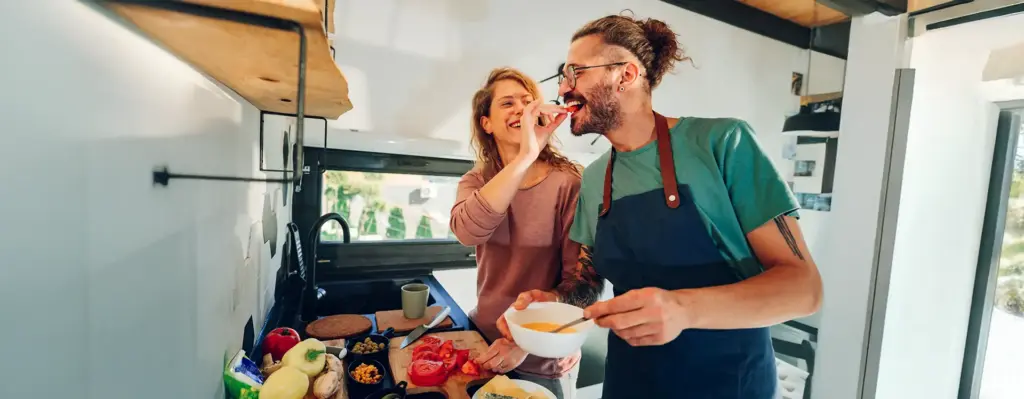 Junges Paar kocht ausgelassen zusammen in der Küche – Frau gibt dem Mann lachend ein Stück Tomate, beide tragen Schürzen.