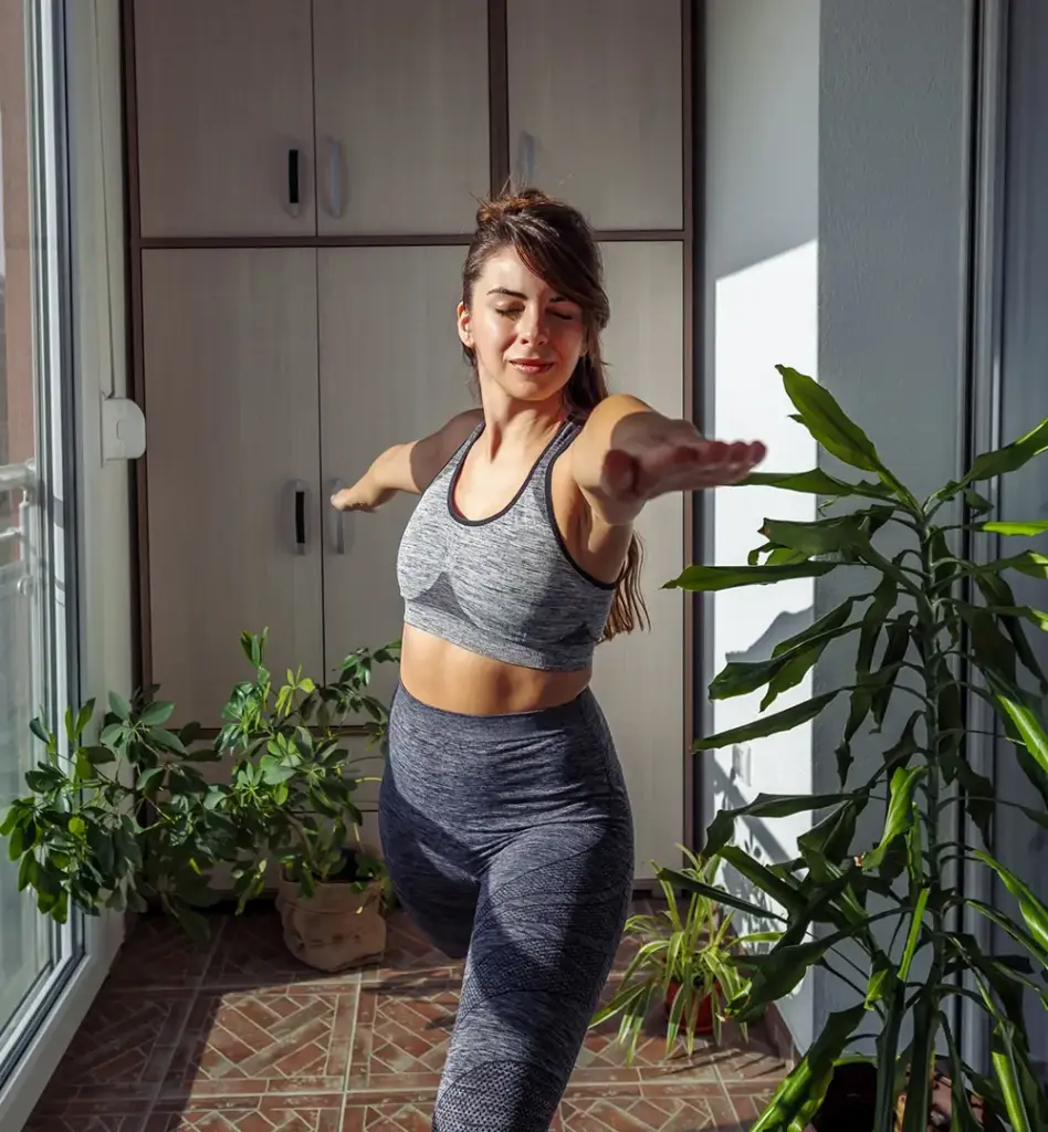 Junge Frau in Sportkleidung macht eine Yoga-Balance-Übung auf einem Bein im hellen Wintergarten – umgeben von Topfpflanzen und Sonnenlicht.