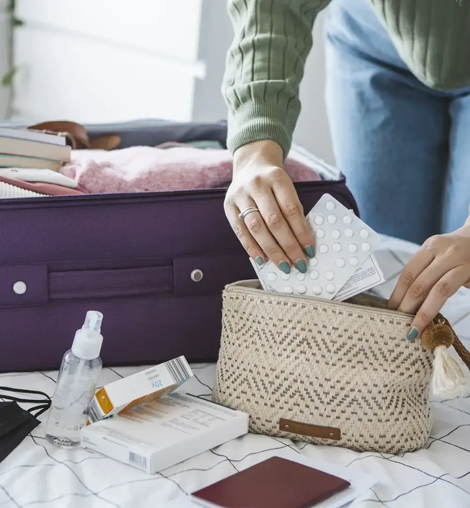 Person legt Medikamente und Tabletten in eine Kulturtasche – geöffneter Koffer und Reisepass im Hintergrund, Vorbereitung der Reiseapotheke.