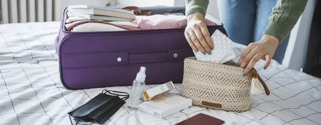 Person legt Medikamente und Tabletten in eine Kulturtasche – geöffneter Koffer und Reisepass im Hintergrund, Vorbereitung der Reiseapotheke.